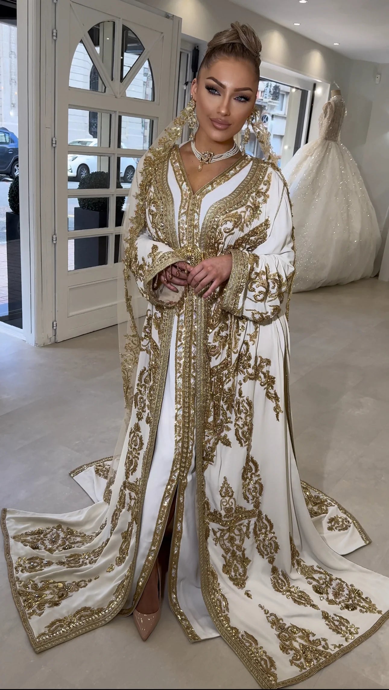 Une femme présente un caftan ivoire orné de détails dorés méticuleux, avec une ceinture bijou captant la lumière. L'étoffe luxueuse dessine une silhouette impériale et gracieuse, évoquant les fastes d'une époque révolue avec une modernité saisissante.