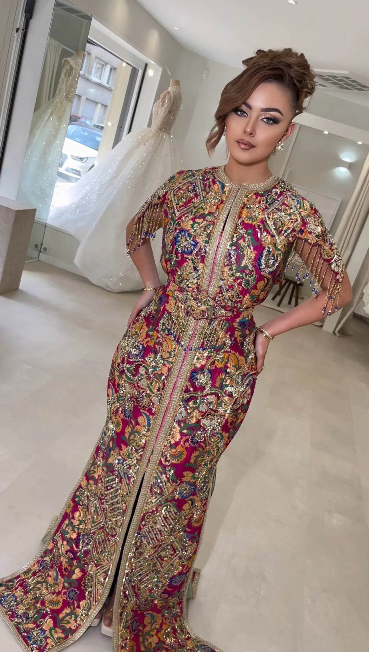 Devant un intérieur lumineux et moderne, une femme se tient avec assurance dans un caftan riche en couleurs et en détails, alliant des tons pourpres et fuchsias avec des broderies dorées étincelantes. Les manches sont ornées de perles qui ajoutent un charme dynamique à la tenue, évoquant les splendeurs d'un jardin fleuri en pleine floraison.