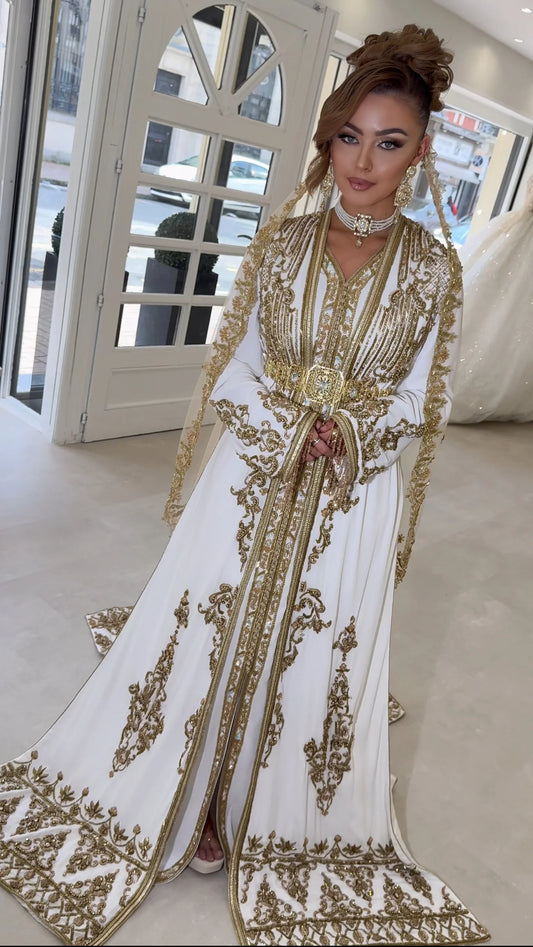 Dans un décor élégant, une femme arbore un caftan blanc orné de motifs brodés en or, évoquant la majesté et le raffinement des palais de l'Alhambra. Sa ceinture, parsemée de pierres précieuses, brille au centre de la tenue, reflétant un goût exquis pour le détail et la tradition.