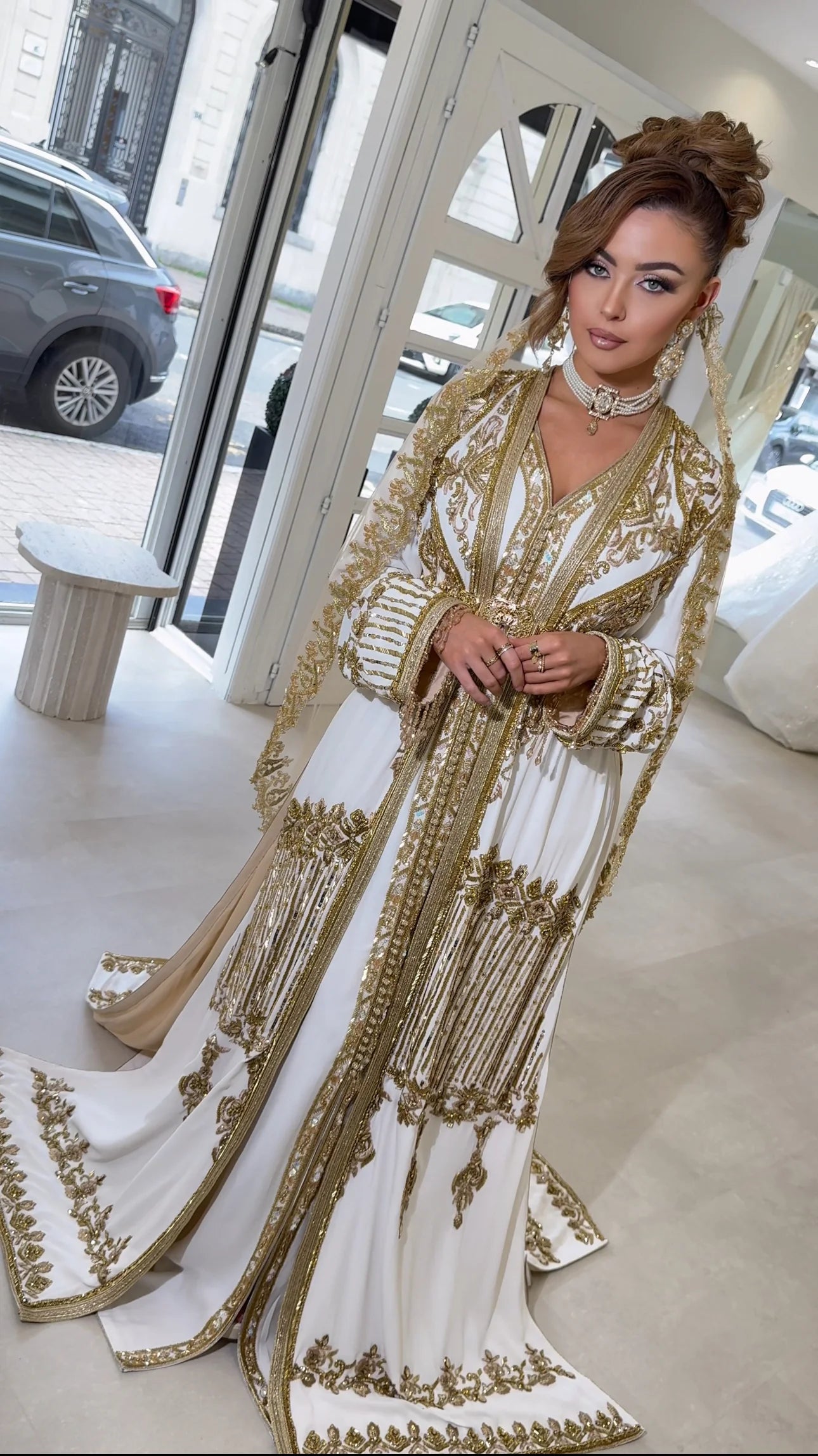 Une élégante femme en caftan blanc orné d'opulentes broderies dorées pose dans un salon de mariage luxueux. Le voile long et richement décoré drapé derrière elle évoque une prestance noble, tandis que les détails de la tenue brillent avec splendeur, reflétant la lumière naturelle de l'espace et l'artisanat raffiné.