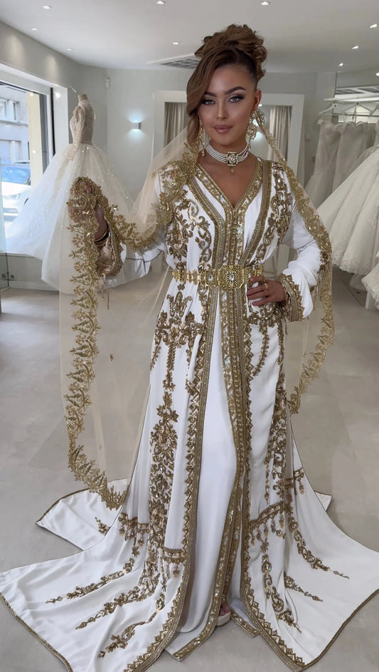 Une femme se tient majestueusement dans un caftan blanc écru orné de broderie dorée complexe, avec une ceinture sertie de pierres brillantes et un voile perlé. L'ensemble dégage une élégance royale, représentant la fusion parfaite de l'artisanat traditionnel et du luxe contemporain.