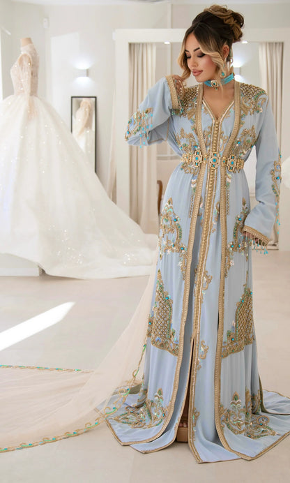 Une femme affiche une allure élégante et sophistiquée dans un caftan bleu ciel pastel avec des broderies dorées et des accents turquoise. Le long voile derrière elle, orné d'une broderie délicate, ajoute une dimension de raffinement et de grâce, tandis qu'elle se tient dans une boutique lumineuse de robes de mariée, incarnant la beauté et l'élégance intemporelles.